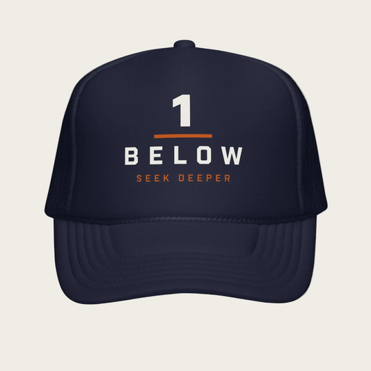 A navy blue trucker hat with a curved brim, featuring a front graphic that reads '1 BELOW SEEK DEEPER' in white and orange text, with mesh side panels.